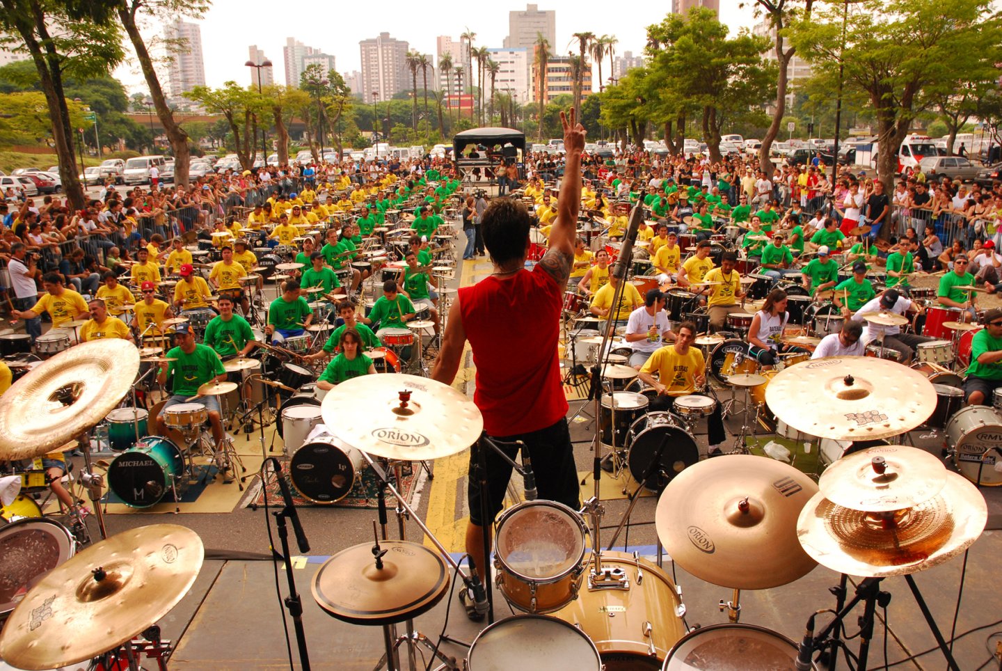 Dino Verdade de costas no palco, braço erguido, regendo centenas de bateristas de camisas verdes e amarelas em uma praça lotada